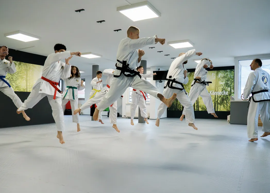 Erwachsene Trainingsgruppe in weißen Doboks springt bei einer Sprung-Tritttechnik synchron ab, ein Trainer beobachtet im Hintergrund