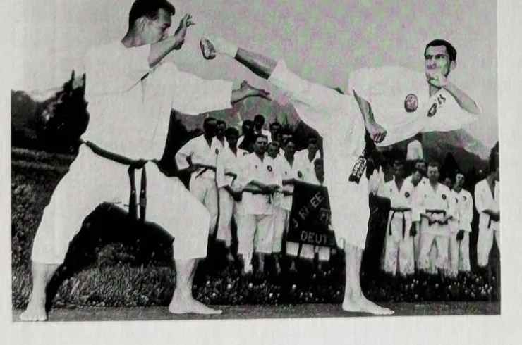 Ein-Schritt-Kampf im Freien: ein Kämpfer führt einen hohen Fußtritt gegen den Partner aus, dahinter steht eine Gruppe in weißen Anzügen vor Bergkulisse, Garmisch 1965