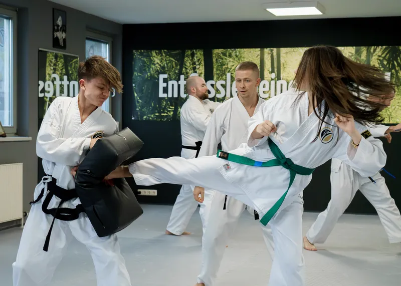 Jugendliche Trainierende in weißen Doboks üben Pratzentraining mit Kicktechniken, im Hintergrund weitere Jugendliche und ein Trainer
