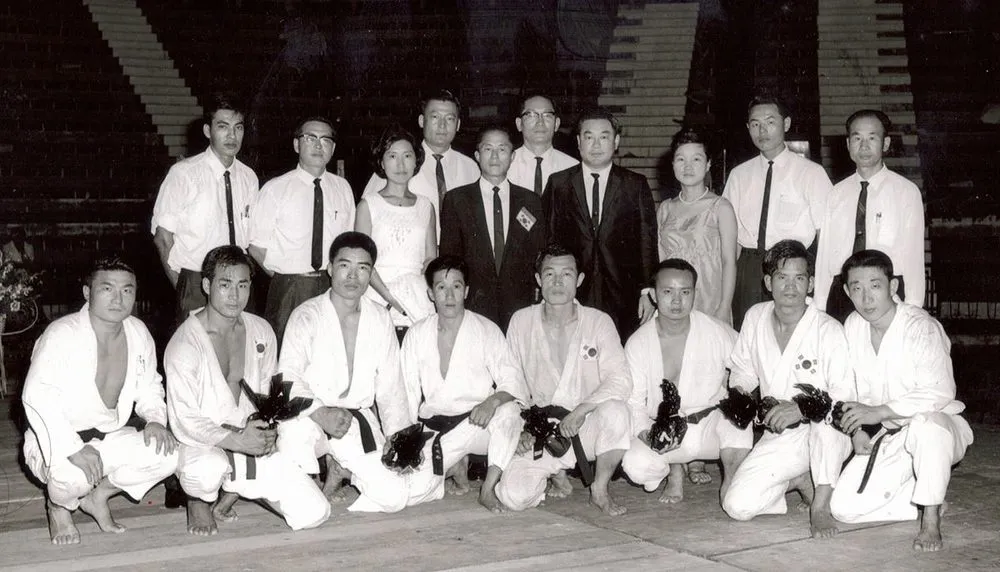 Gruppenbild in einer Halle: Männer im Dobok mit Blumensträußen kniend, dahinter Funktionäre in Anzügen
