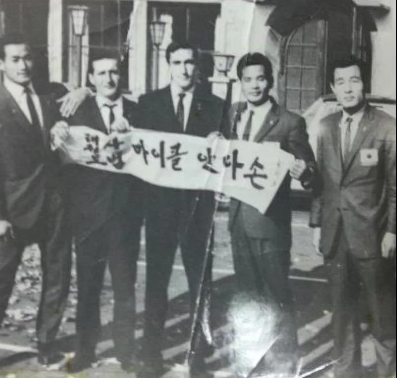 Mike Anderson im Dobok hält mit zwei Begleitern ein koreanisches Transparent, Dezember 1964