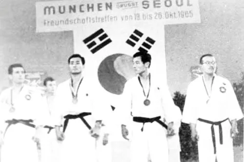 Männer im Dobok mit Medaillen unter dem Banner „München grüßt Seoul – Freundschaftstreffen vom 19. bis 26. Okt. 1965“ und koreanischer Flagge