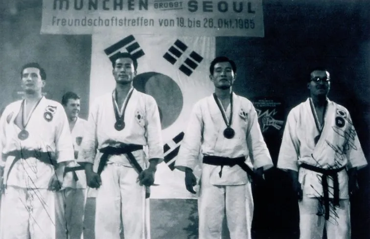 Vier Männer im Dobok mit Medaillen vor koreanischer Flagge und dem Banner „München grüßt Seoul“