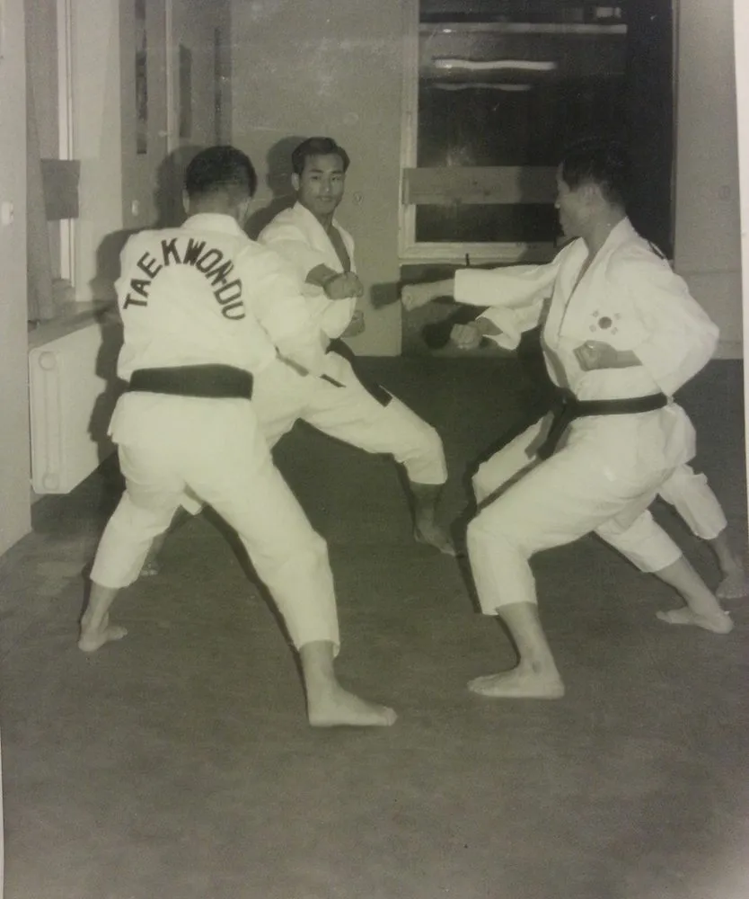 Zwei Männer im Dobok bei einer Partnerübung in einer Halle, einer mit Rückenaufschrift Taekwon-Do