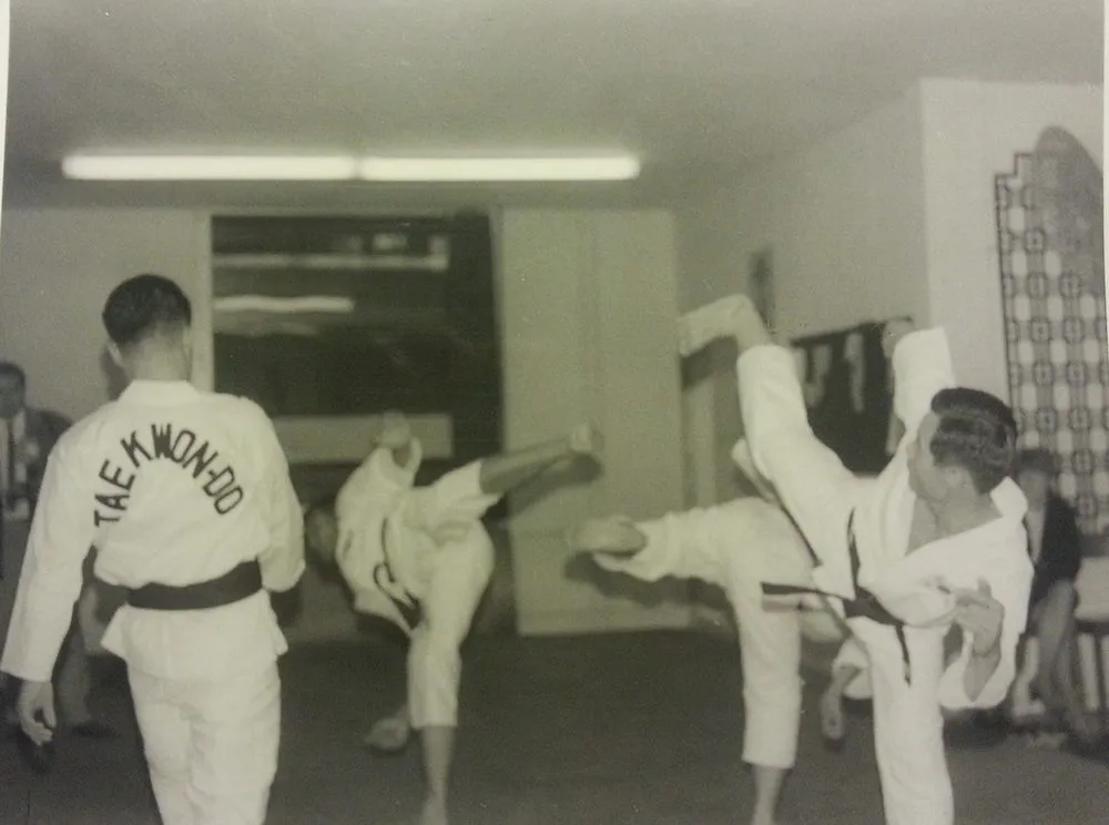 Ein Mann im Dobok führt einen hohen Sprungtritt aus, ein Partner mit Rückenaufschrift Taekwon-Do steht daneben
