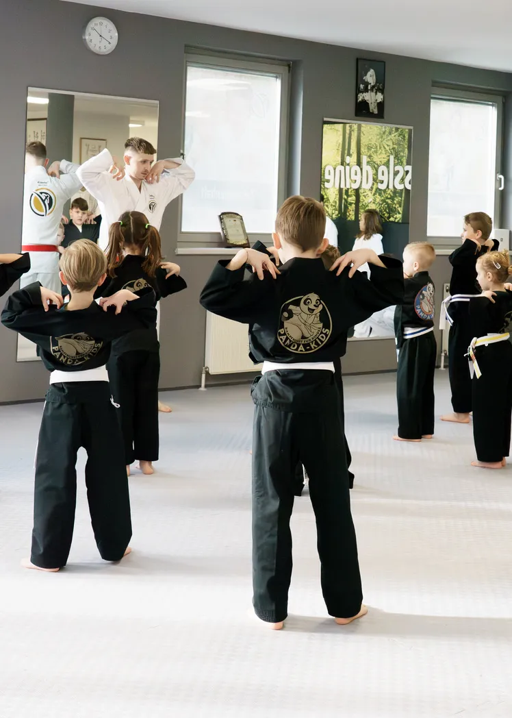 Kindergartenkinder in schwarzen Panda-Kids-Trainingsshirts stehen mit dem Rücken zur Kamera und dehnen sich, ein Trainer macht die Übung vor