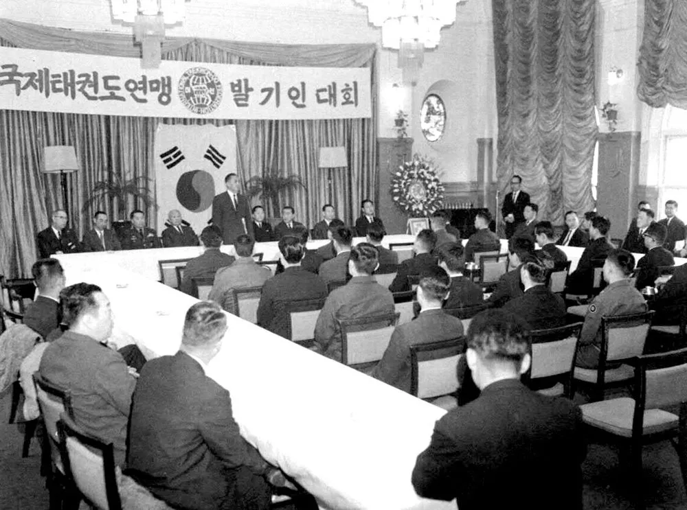 Sitzung an langen Tischen in einem Saal, an der Stirnseite ein koreanisches Transparent und die Nationalflagge