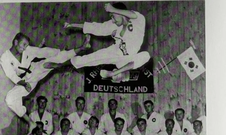 Zwei Taekwondo-Kämpfer in Sprungtechniken vor einer Gruppe sitzender Schüler, im Hintergrund ein Banner „J. Rhee Inst. Deutschland“ und die koreanische Flagge, Garmisch 1965