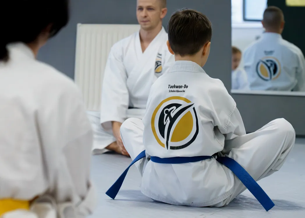 Grundschulkind im weißen Dobok mit blauem Gurt sitzt im Schneidersitz aufmerksam vor einem sitzenden Trainer